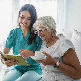 Woman reading a book while sitting with happy grandmother drinking tea. Happy elder woman sitting on white sofa and listening to nurse reading a book out loud